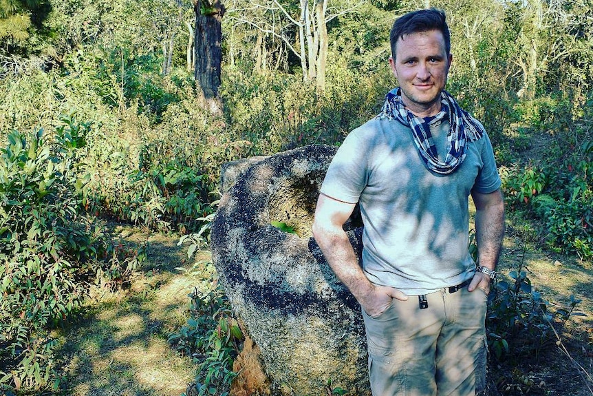 A man leans against a large stone jar in the jungle