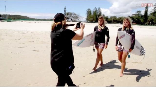 Rear view of woman using small camera to film two girls walking with surfboards on the beach