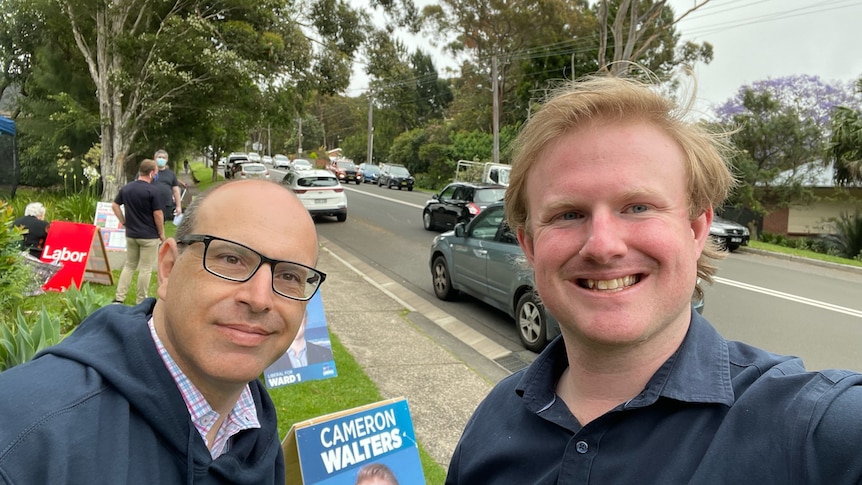 Two men take a selfie in front of a campaign sign