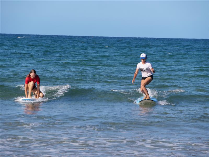 Dos mujeres en el agua, una arrodillada sobre su tabla y la otra parada sobre su tabla montando una pequeña ola.