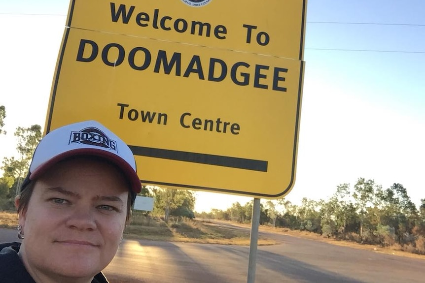 A woman in a cap stands in front of a yellow sign saying "Welcome to Doomadgee town centre "in black writing.