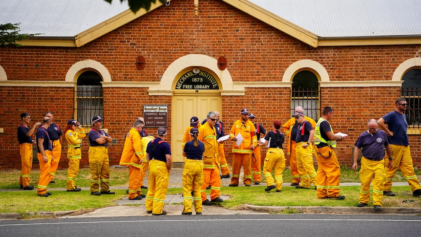 Firefighters in bright orange uniforms gathering outside a brick building.