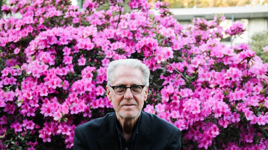 Garth Popple sits on a wall in front of a bush of bright pink flowers