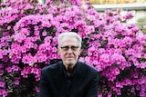 Garth Popple sits on a wall in front of a bush of bright pink flowers