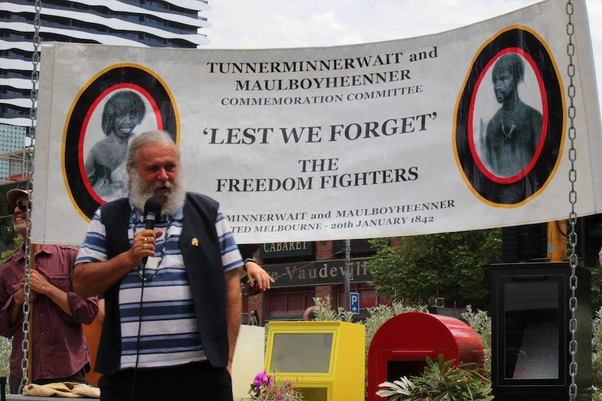 A bearded man speaks in front of a banner which reads "lest we forget the freedom fighters".