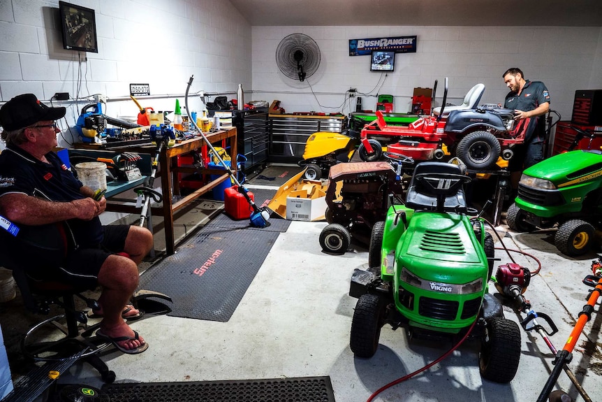 A man repairs a ride on mower while another man watches on.