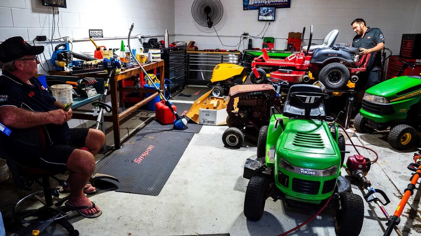 A man repairs a ride on mower while another man watches on.