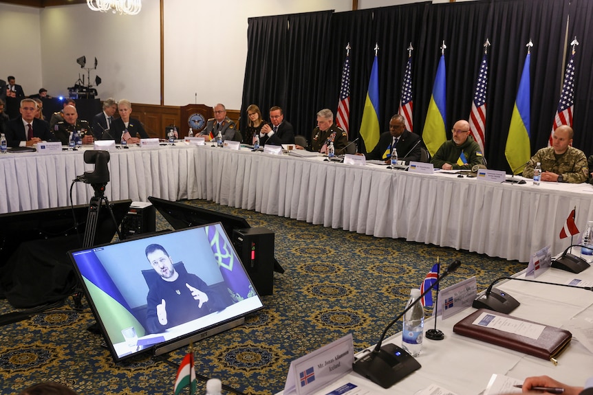 Defence leaders seated around table look onto screen showing Volodymyr Zelenskyy speaking.