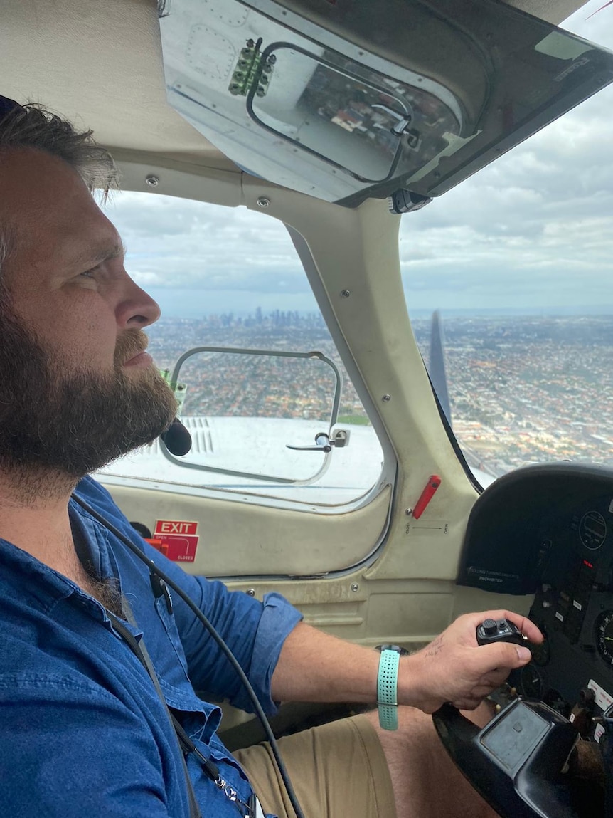 A blonde man with a beard and hands on a steering wheel looks out of a helicopter window.