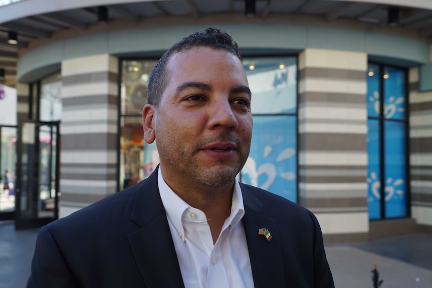 Headshot of Jason Wells who wears a suit and stands in front of shops