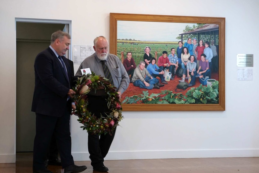 Two men wearing suits and ties solemnly carry a floral wreath in front of a colourful painting of 15 people on a farm