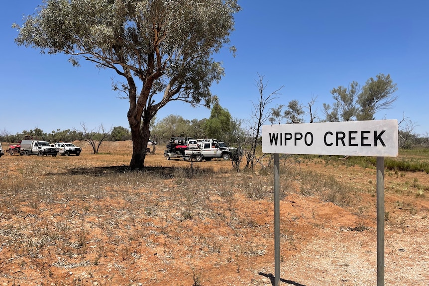 A sign reading Wippo Creek stands on bare ground amid sparce, low scrub with four-wheel drive vehicles in the background.