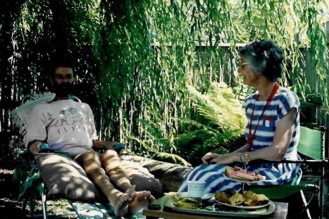 A thin, gaunt man with dark facial hair sits in a chair in a garden, with a grey-haired woman in a striped dress beside him.