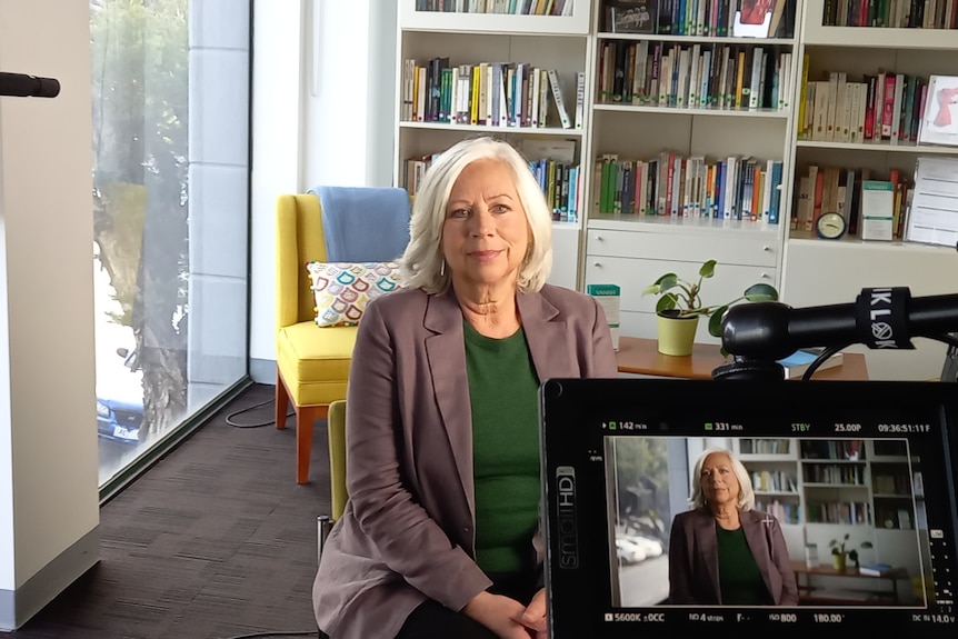 A woman with shoulder length silver hair and a mauve blazer sits in an office surrounded by camera equipment