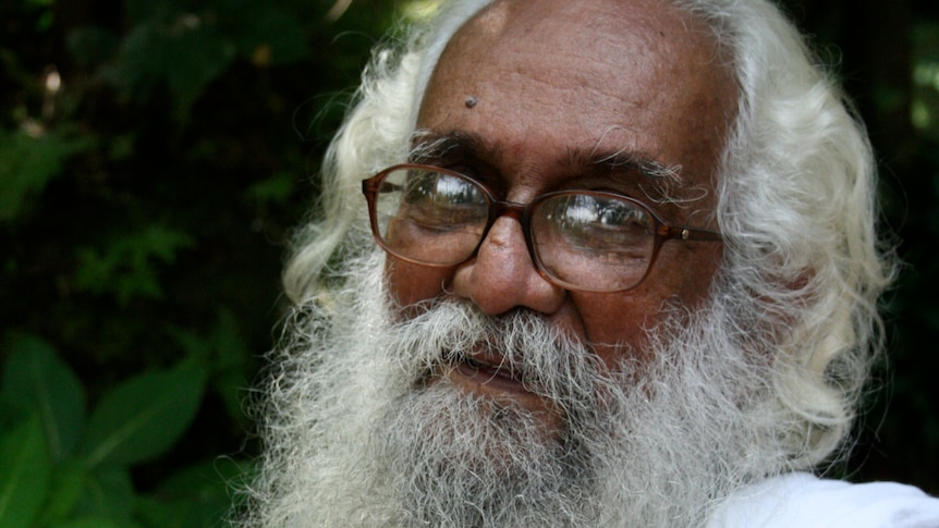 A man with long white hair and white beard, with glasses, smiles at the camera.