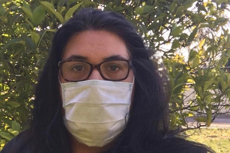 A woman with long dark hair and glasses wears a face mask.