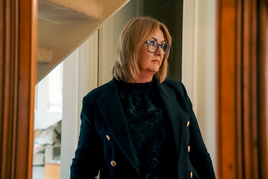 Jenni wears glasses, a black brass button blazer over a dark pattereed blouse and stands under a staircase looking off camera