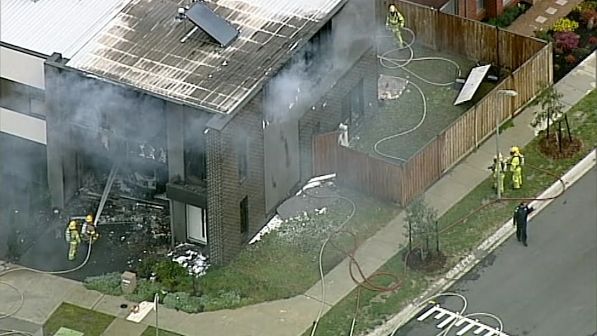An aerial view of a townhouse blackened by fire, surrounded by smoke and firefighters in bright yellow uniforms.