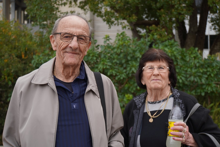 An older couple stand for a photo with the woman on the right holding a bottle of soft drink.