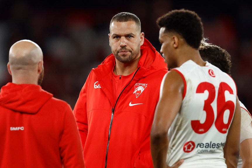 Sydney Swans star Lance Franklin stands wearing a team jacket as he talks to teammates and an assistant coach during a game.