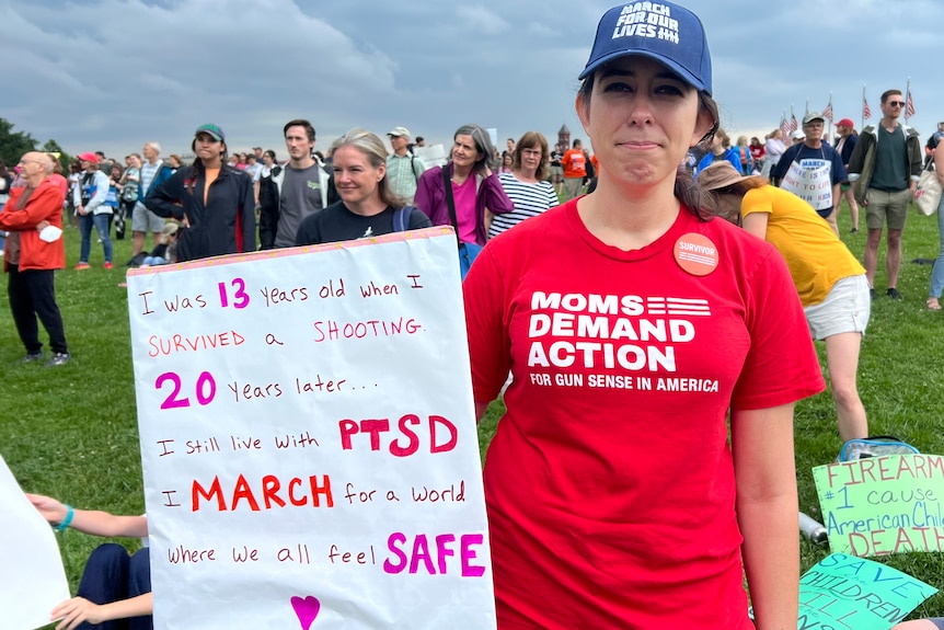 Cristina Little in a red shirt reading moms demand action and holding a placard detailing the PTSD she has suffered