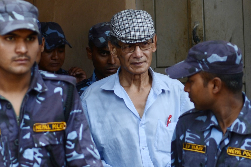 Charles Sobhraj wearing handcuffs and escorted by police out of a building.