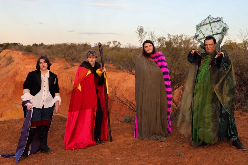 Four people in bright costumes stand on red dirt with a blue sky behind them.