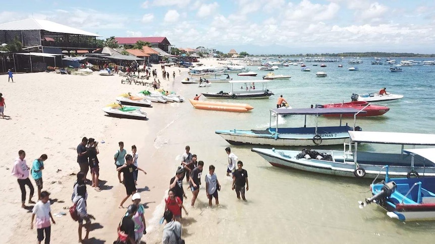 People play on the beach in Bali while boats sit on the white sand.
