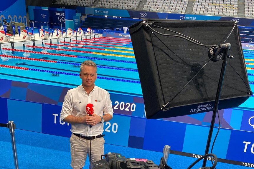 A TV reporter stands holding a microphone with "7" on it, next to a swimming pool at the Tokyo Olympics.