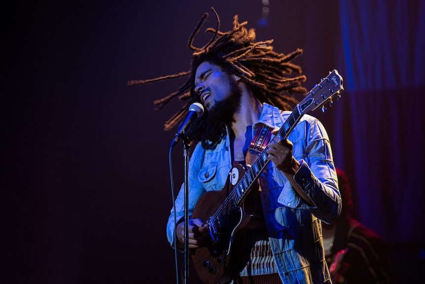 Kingsley Ben-Adir as Bob Marley holding a guitar, shaken hair, singing into a microphone, blue tinge to photo