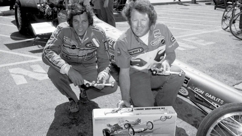 Two racecar drivers in uniform squat while holding toy Hot Wheels cars