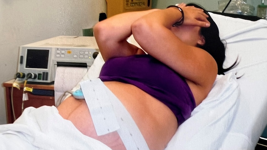A woman who has given birth rests her hands on her face as she lies in a hospital bed with monitoring equipment on her belly.