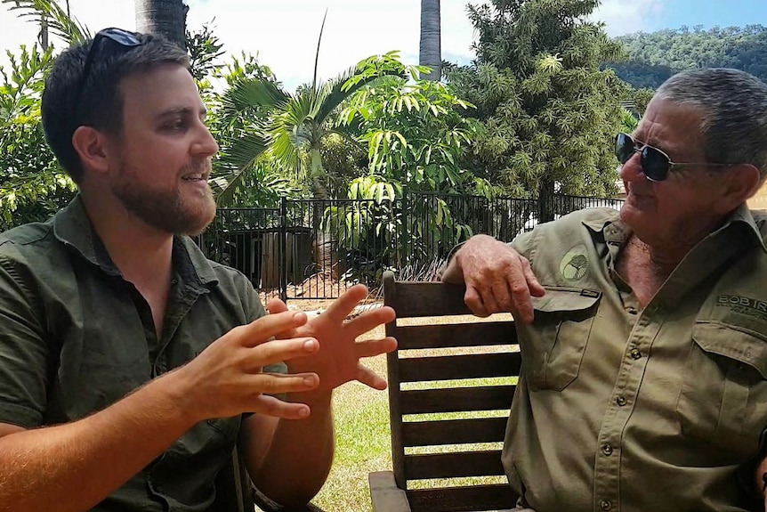 Two men wearing green and khaki shirts speak in a tropical garden.