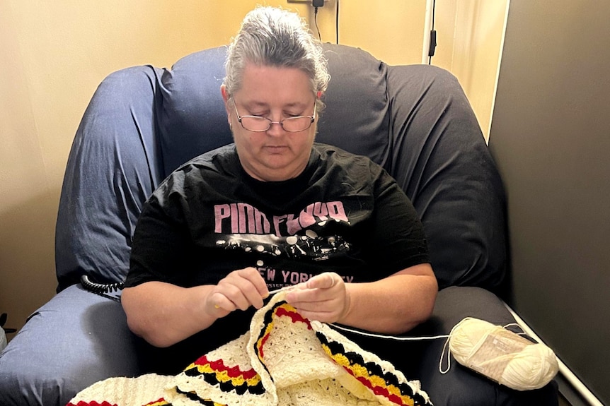 A woman wearing glasses sitting in a chair making a crochet blanket