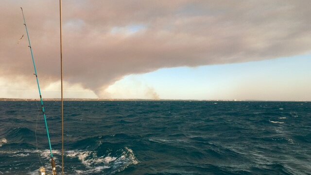 Thick grey smoke from a bushfire fills the sky over the ocean off Perth's coast, with a fishing rod on the front of a boat.