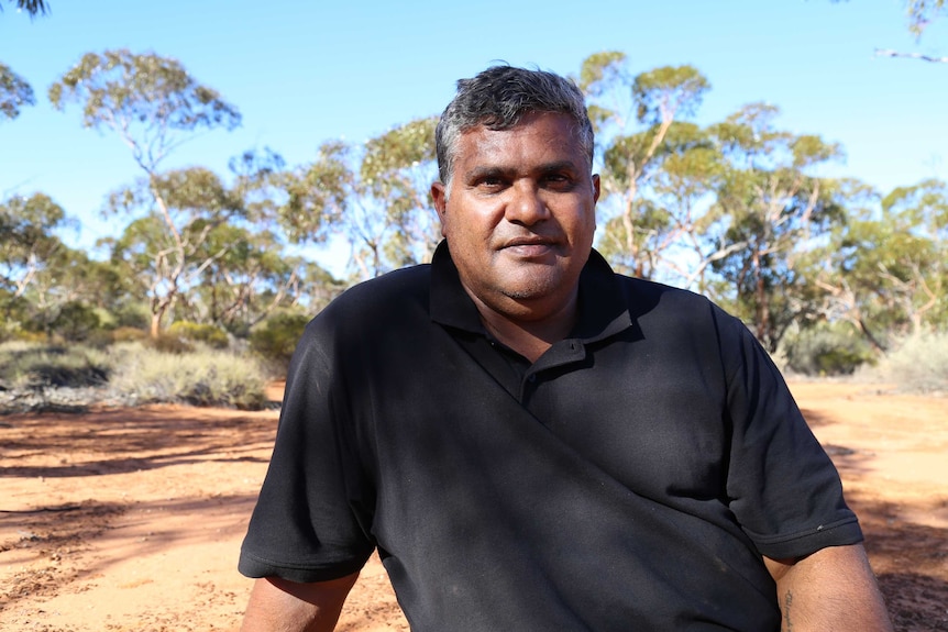 An Indigenous man in the foreground with trees and blue sky in the background.