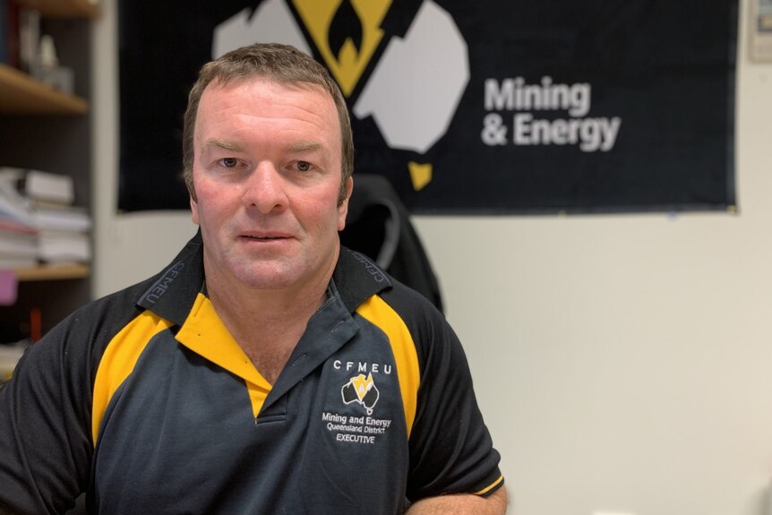A man in a polo shirt. He has brown hair. There is a mining and energy sign in black, white and yellow behind him.