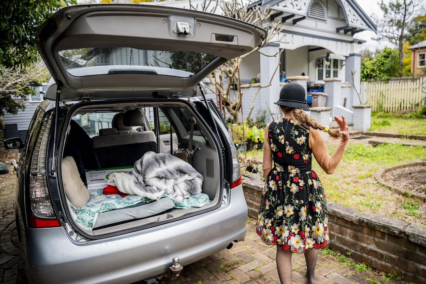 A woman is seen from behind in a dress and hat, standing by a car with the back door open, showing a pillow and a blanket.