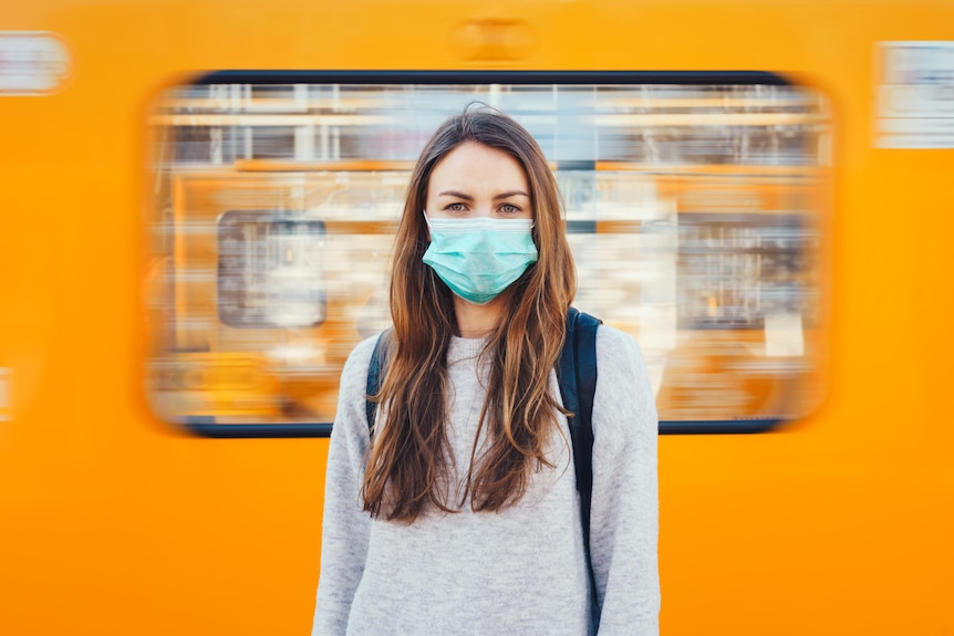 A woman wearing a surgical mask stands in front of the moving train.