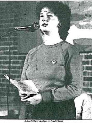 Black and white image of a woman standing at a microphone wearing a jumper and dark trousers