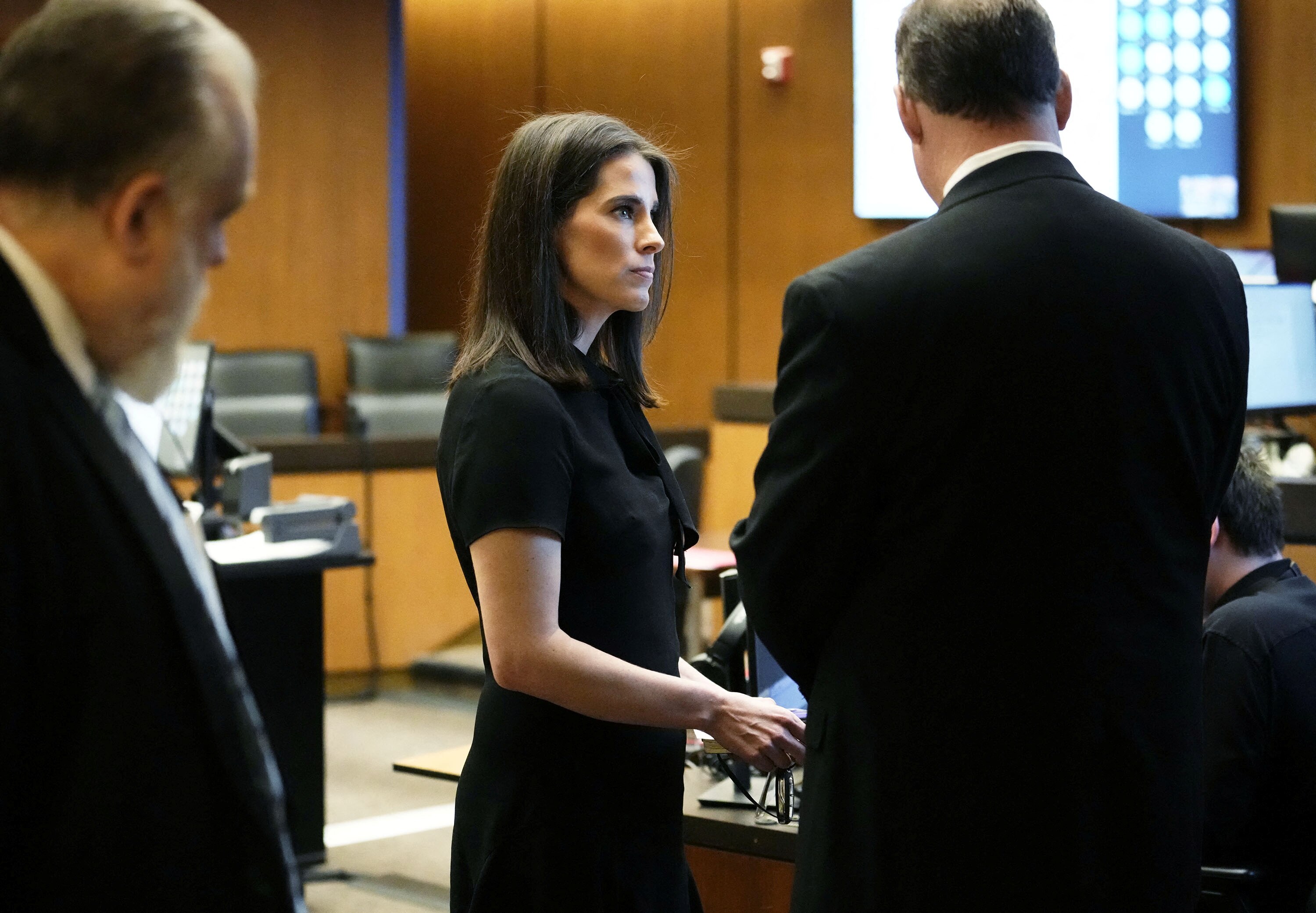 Uma mulher vestida de preto está entre homens de terno em um tribunal.