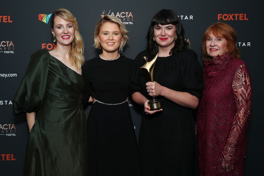 Four women in ball gowns pose in front of a press screen emblazoned with AACTA AWARDS and FOXTEL. One holds a gold statue