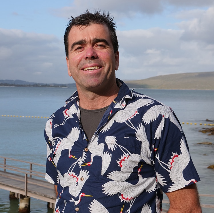 A smiling Andrew Collins with calm waters and a timber jetty in the background.