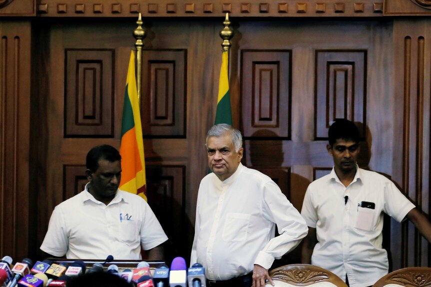 Ousted Sri Lankan PM Ranil Wickremesinghe approaches a row of microphones with two Sri Lankan flags behind him.