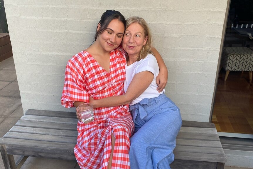 A young woman wearing a red gingham dress sits on a bench beside her mum. They have their arms around each other and smile. 