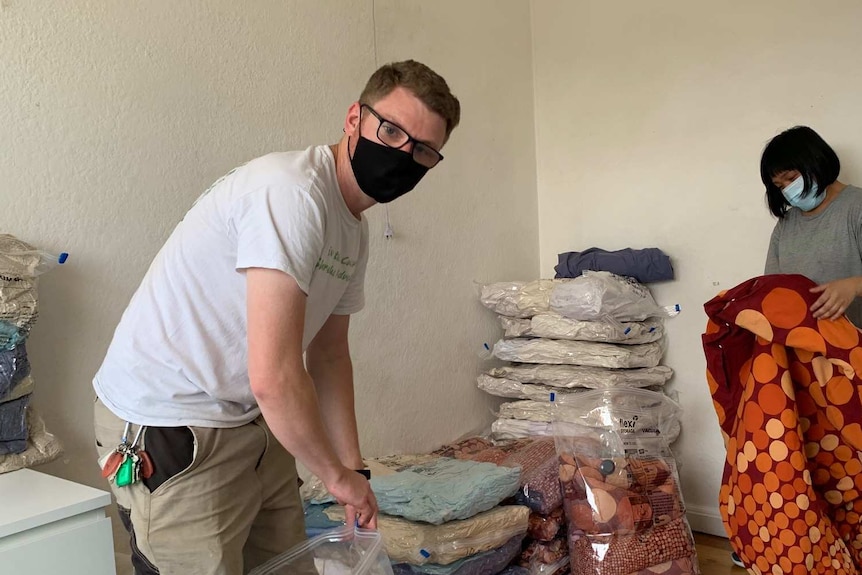 A man packing bags with a woman in the background, both wearing covid safe masks.