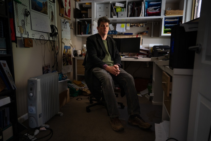 A man sits in a shadowy office and looks at the camera.