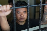 Man inside Kerobokan prison grips onto the bars and looks into the camera.