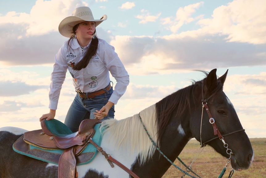 Tyler Morton lifting herself onto a black and white horse wearing jeans, white long sleeve top and a hat.
