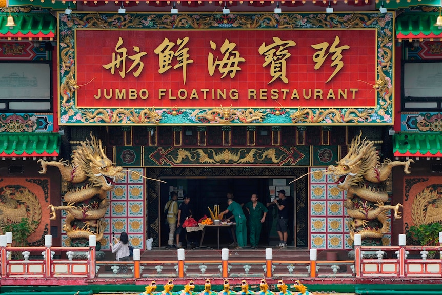 People gather at a table at the entrance to a restaurant flanked by two statues of giant dragons 